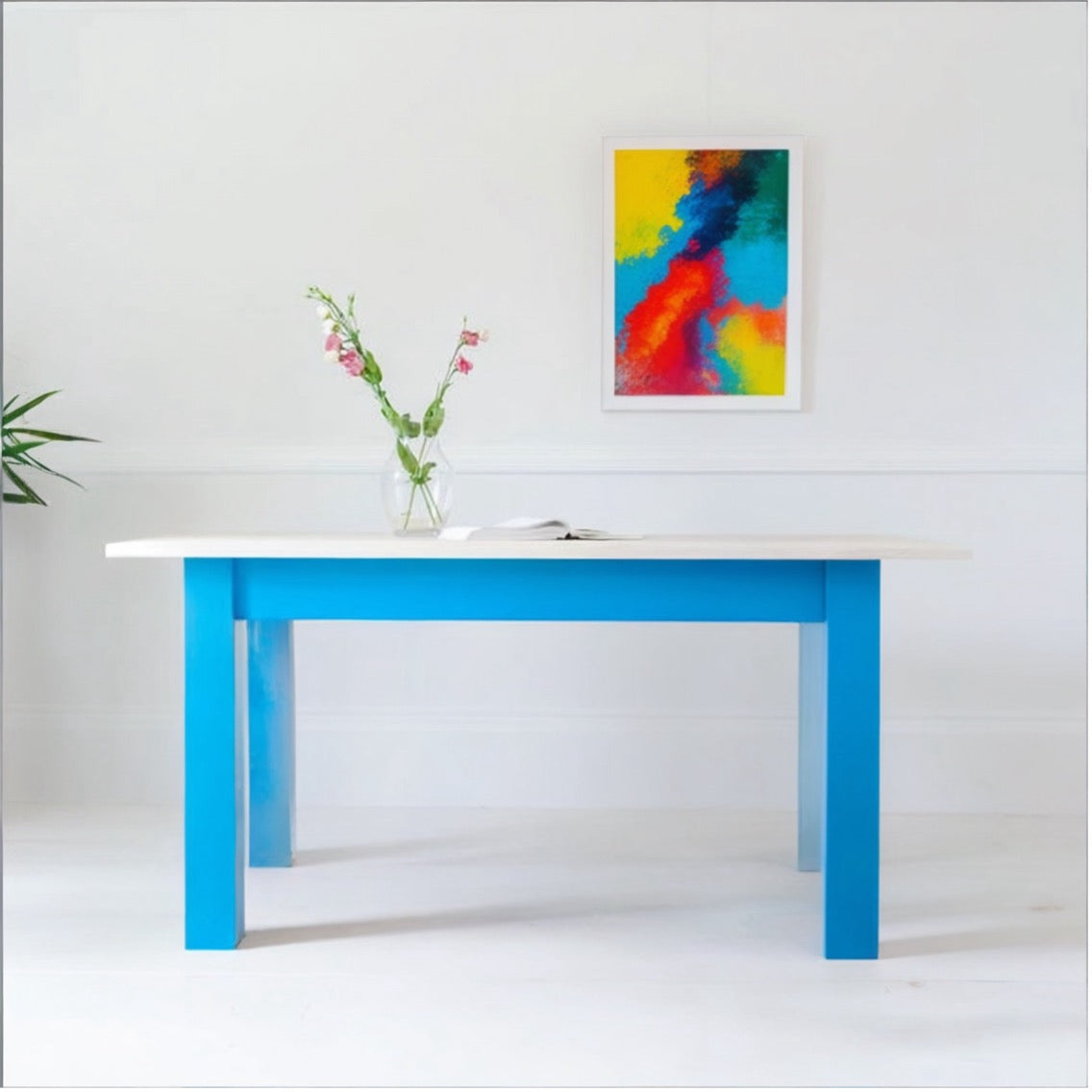 Wooden Farmhouse table with blue legs in a white room with picture on the wall.